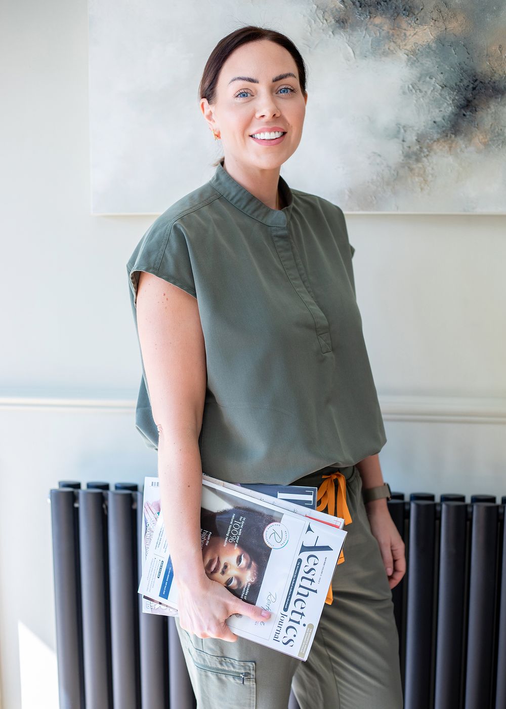 Someone in an olive green dress holds a stack of books while standing against a gray textured background.