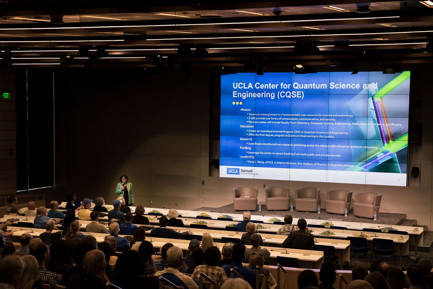 Conference attendees seated in a lecture hall with a large blue presentation screen displaying UCLA content.