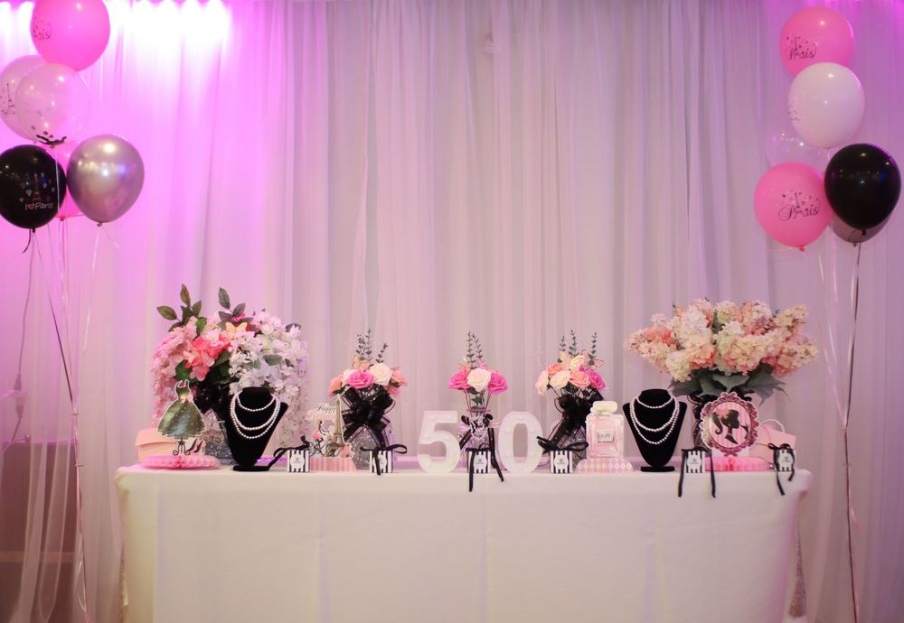 Pink and black balloon decorations with floral centerpieces create an elegant party setup against white draped backdrop.