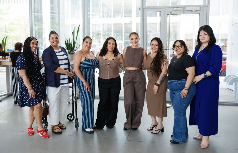 Group of eight diverse women smiling together indoors at a bright, modern venue for a social gathering.