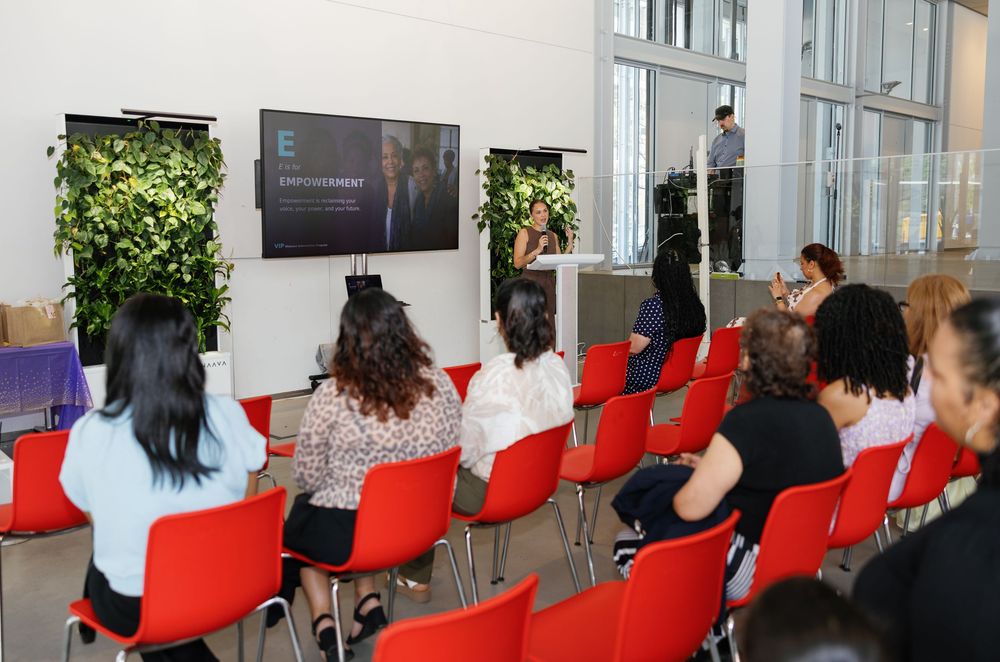 Audience seated in red chairs at a corporate presentation with a speaker at podium and green wall backdrop.