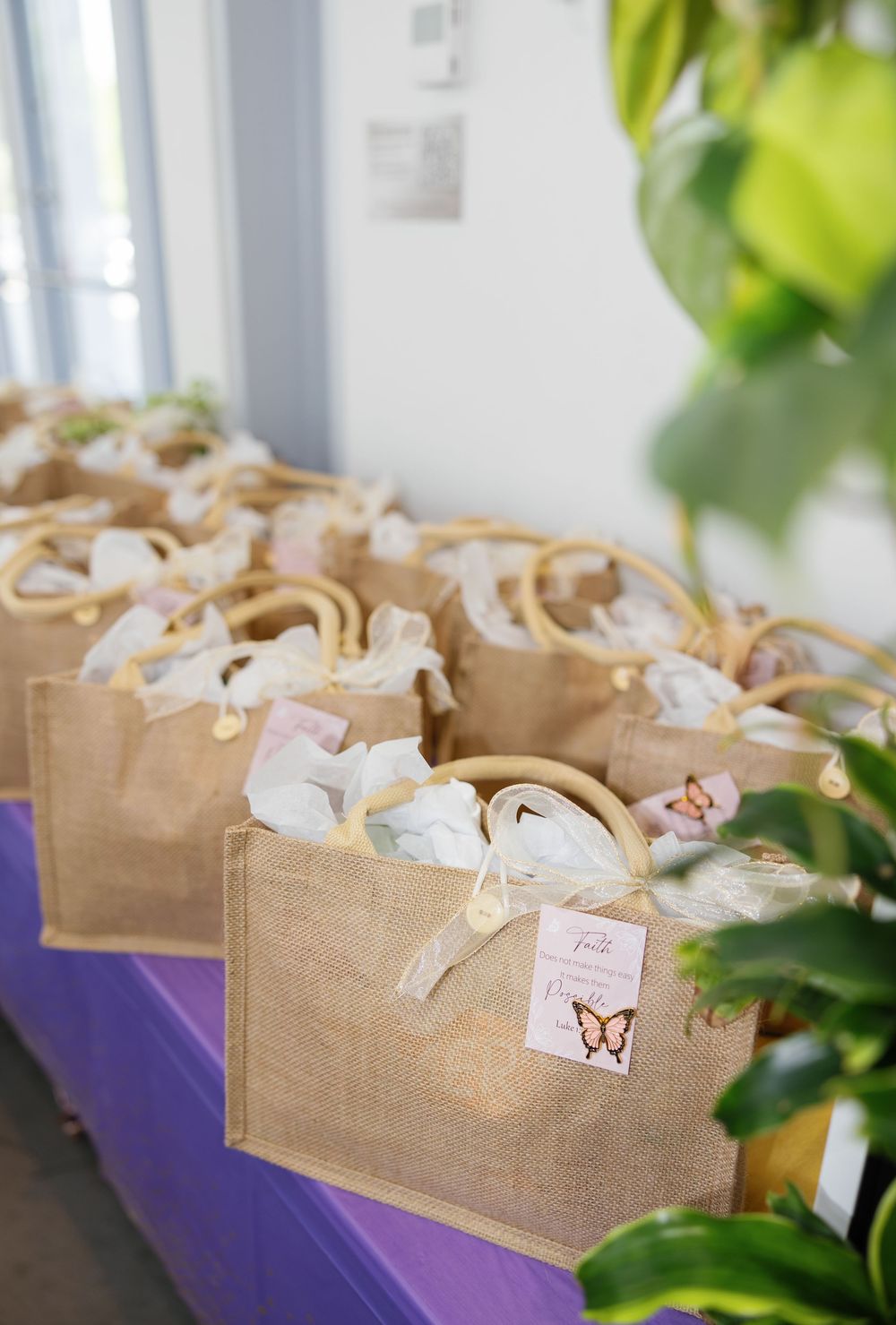 Burlap gift bags with ribbon bows and butterfly tags arranged on a purple table for a party or wedding.