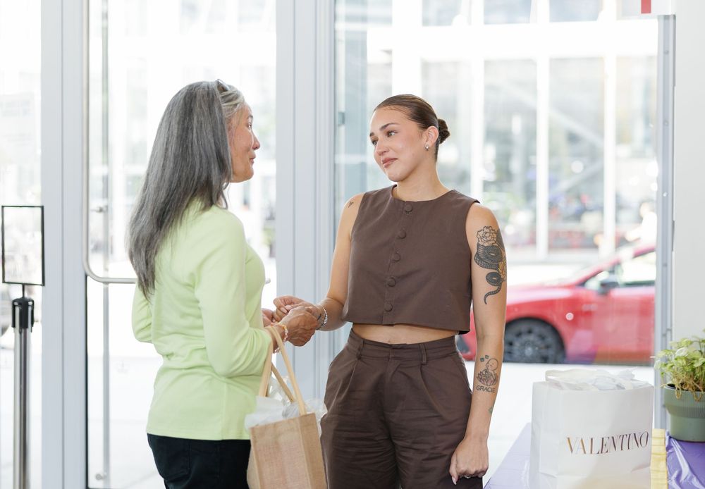 Two women chat near a Valentino store entrance, one with a tattoo wearing brown coordinates, the other in a green jacket.