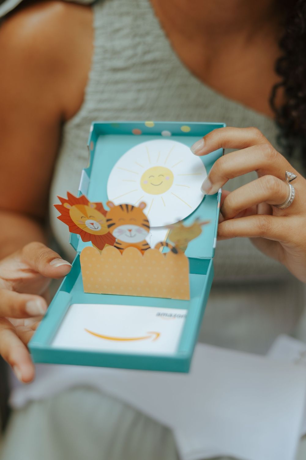 Woman holding an open Amazon gift card box with cute jungle animal pop-up design and smiley sun decoration.