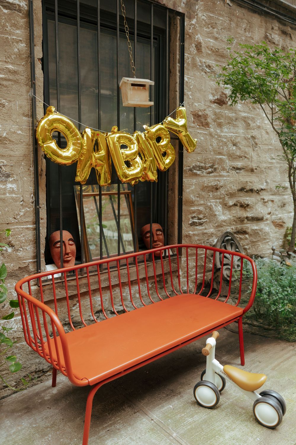 Red metal bench with gold BABY balloon letters and decorative masks against a stone wall with a small tricycle nearby.