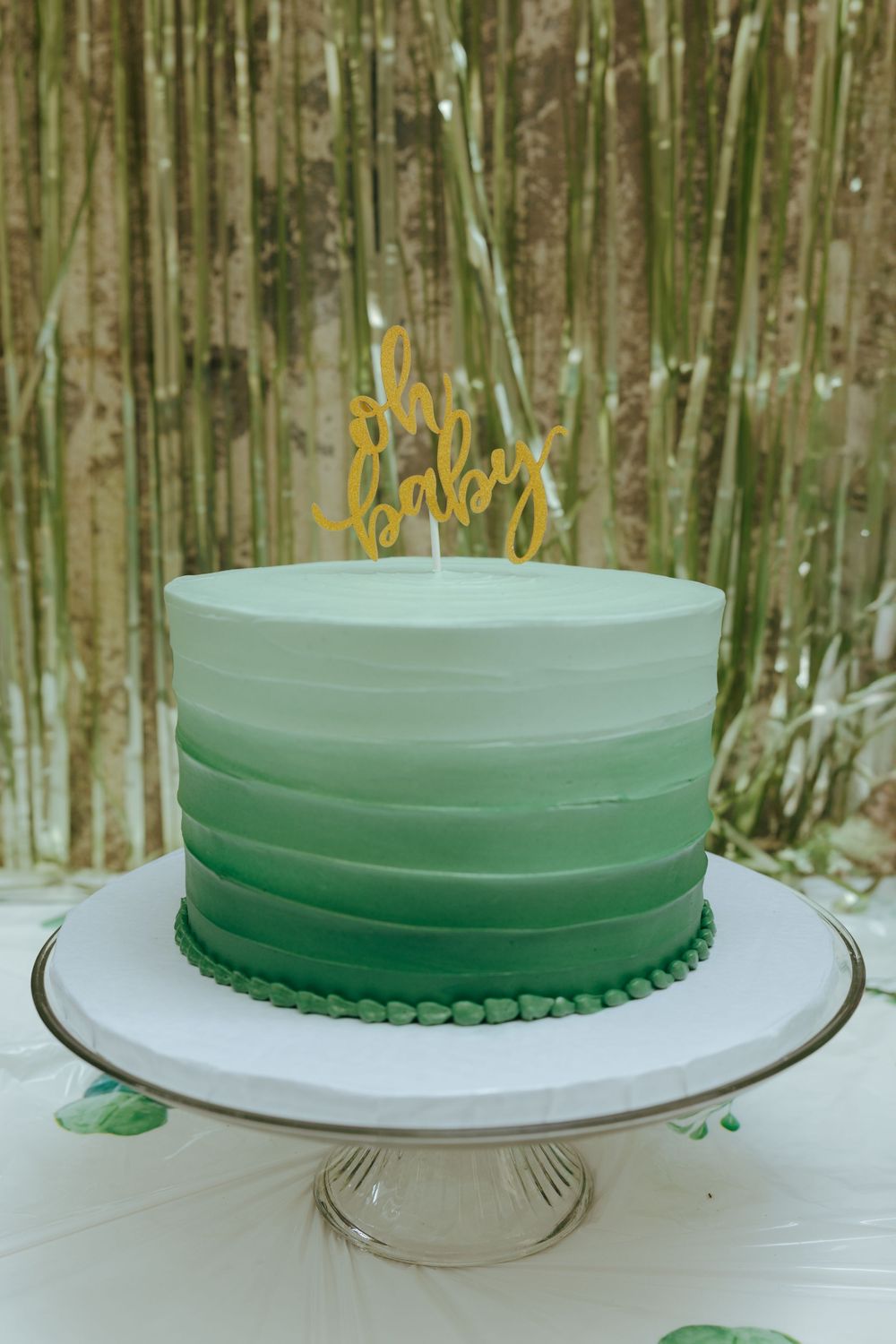 Green ombre baby shower cake with gold oh baby topper on white cake stand outdoors with bamboo background.