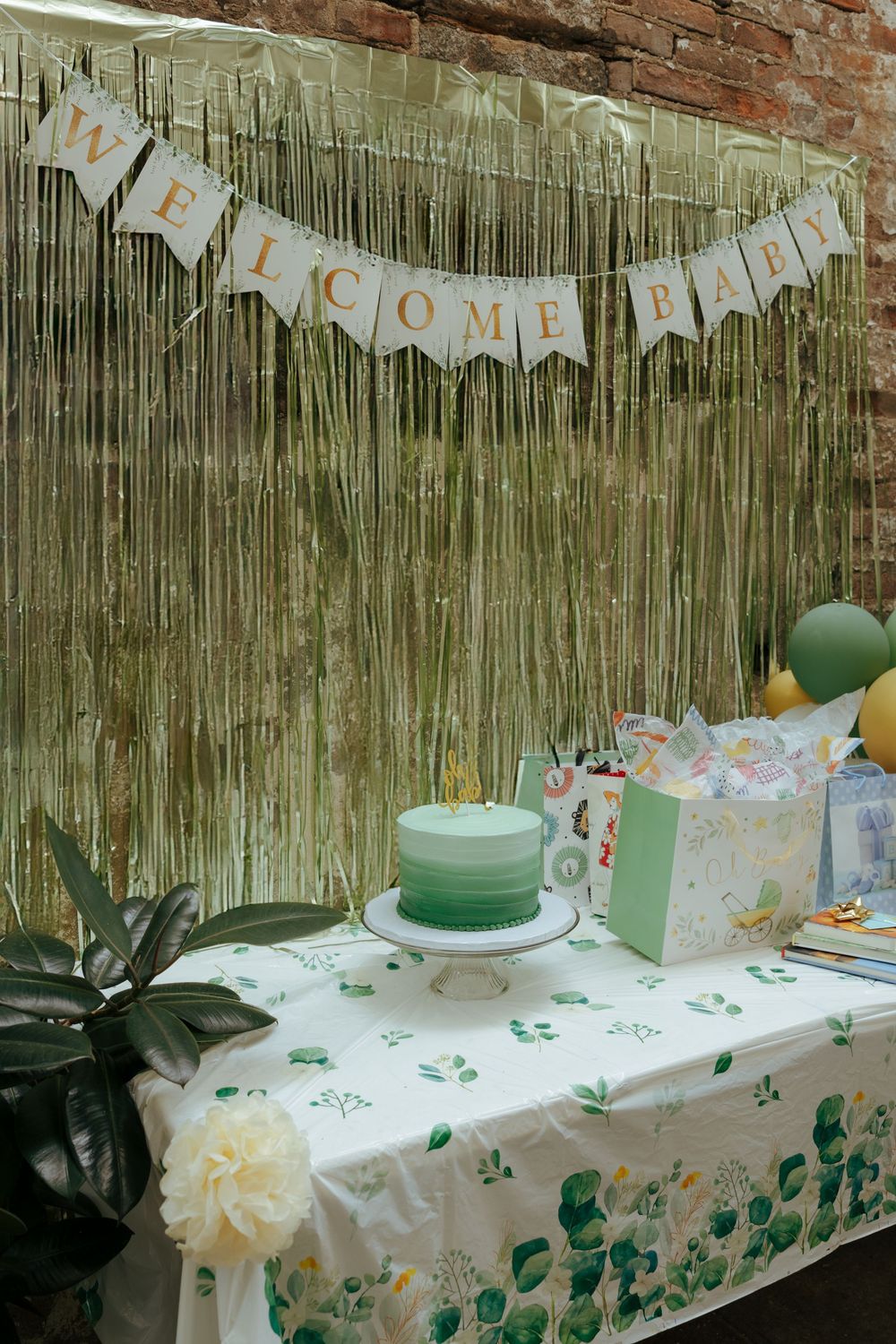 Welcome Baby shower table with green ombre cake, gold tinsel backdrop, banner, balloons, and floral tablecloth decorations.
