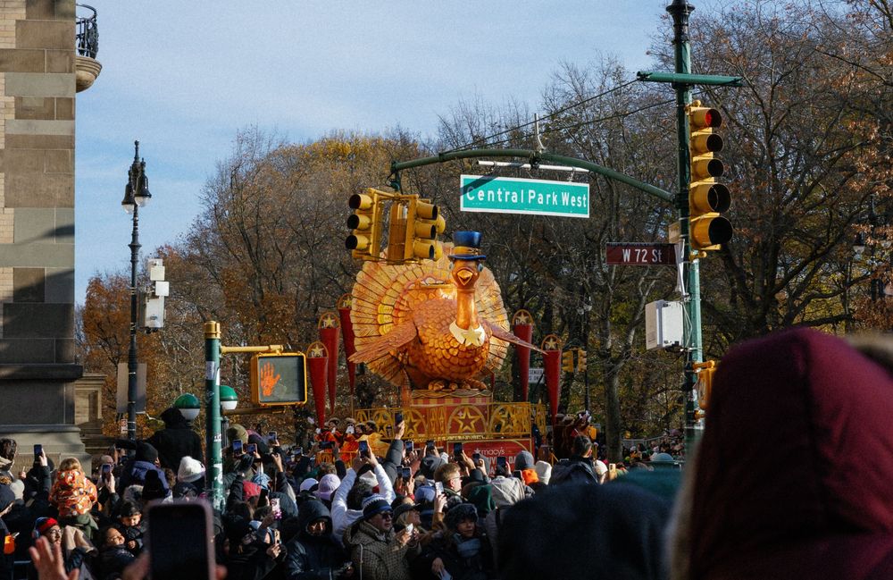 A panoramic view of a busy city street corner with autumn trees, lamp posts and pedestrians waiting to cross.