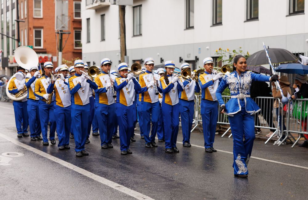 Marching band members in blue and white uniforms perform while walking down a city street during a parade.