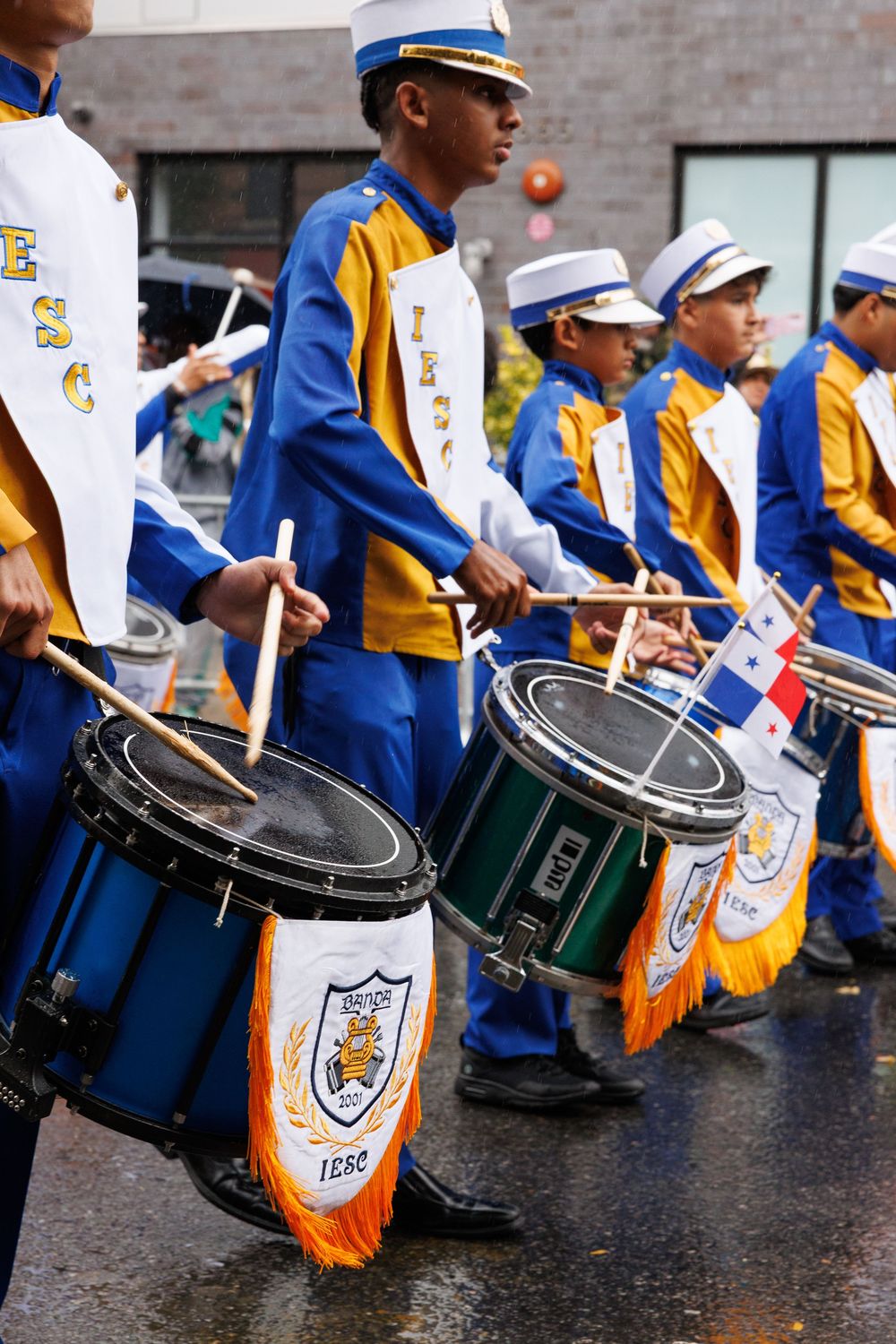 Marching band drummers in blue and gold uniforms perform with snare drums during a parade on wet pavement.