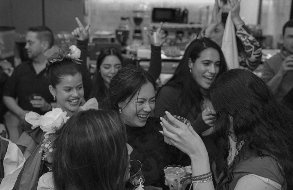 A candid black and white photograph shows people laughing and enjoying themselves at a social gathering.