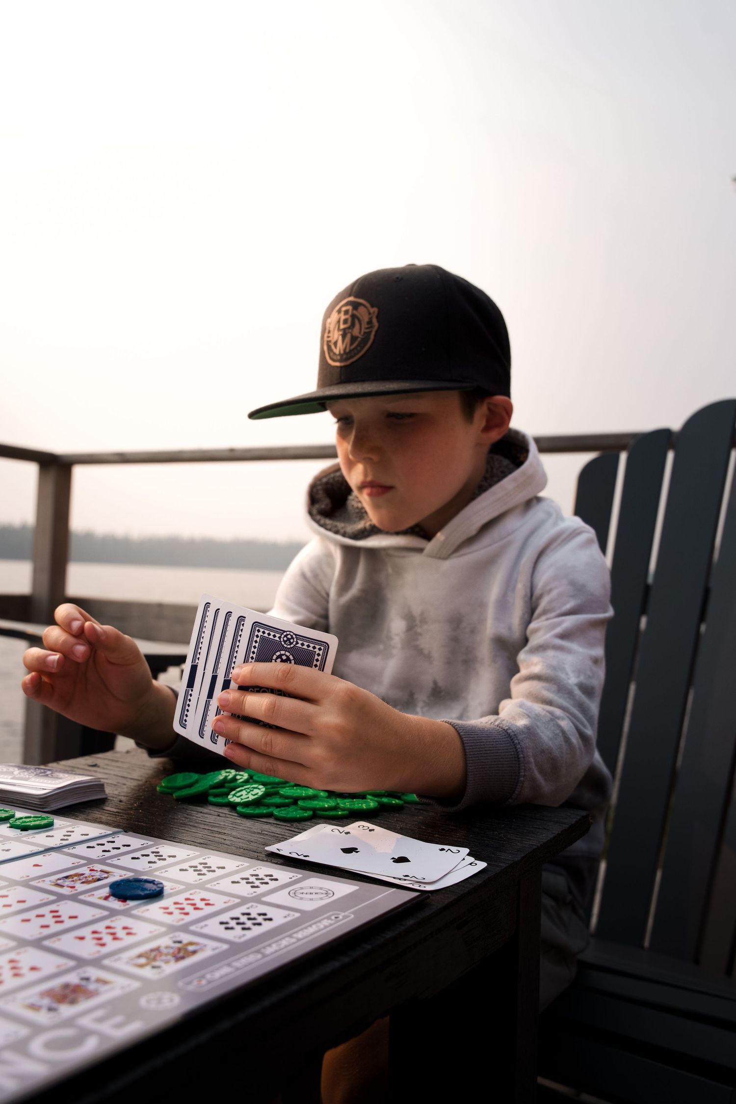 A card player wearing a baseball cap and hoodie sits at a table with playing cards and poker chips.