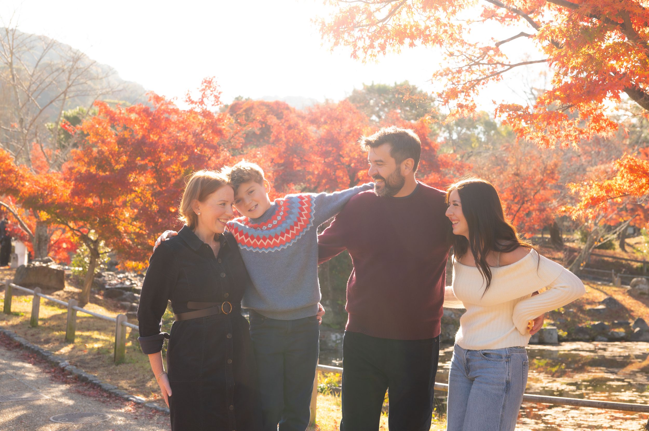 A family of four laughs together outdoors surrounded by brilliant red autumn foliage.