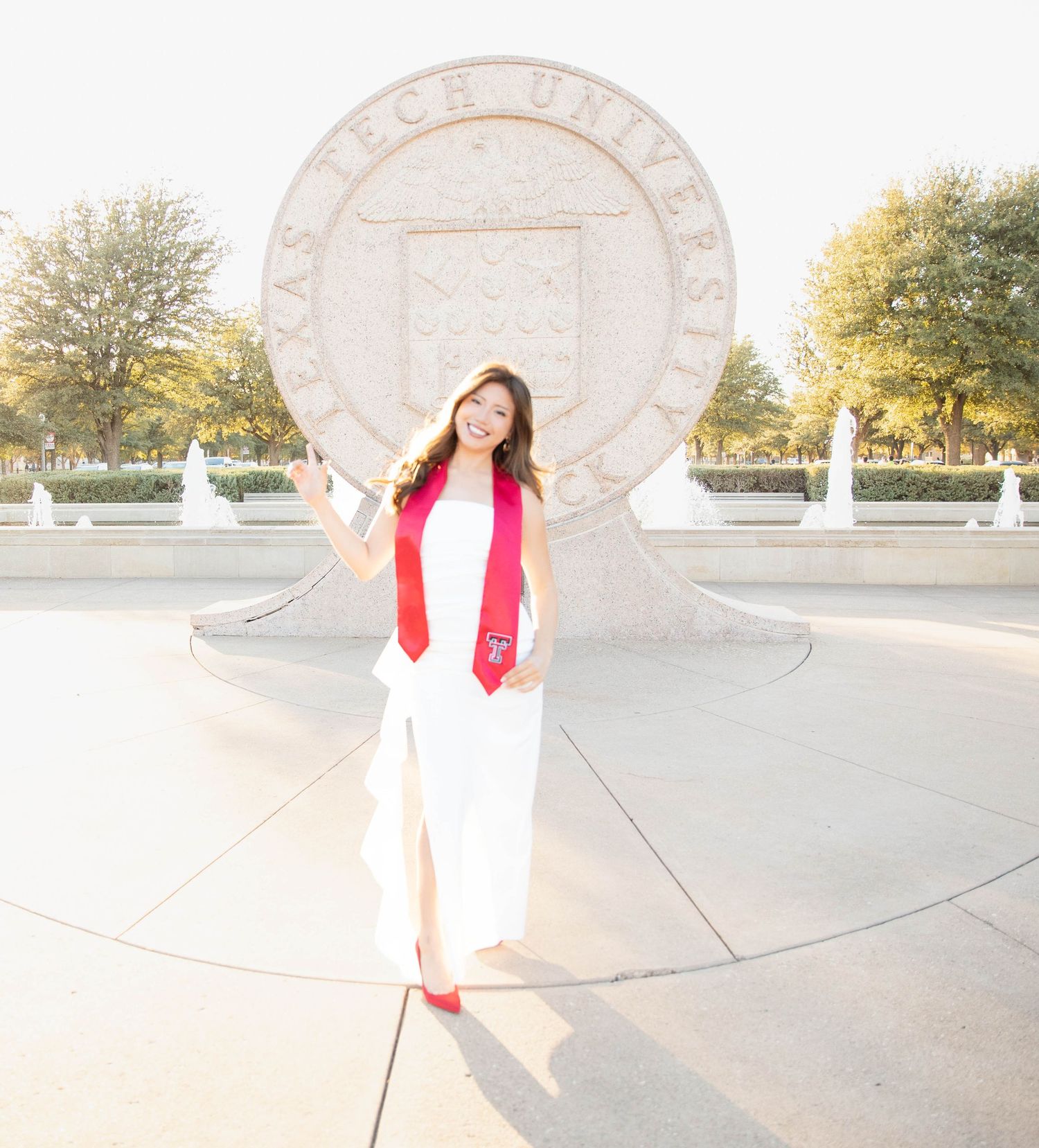 Graduate in white dress and red stole poses in front of large university seal on sunny day.
