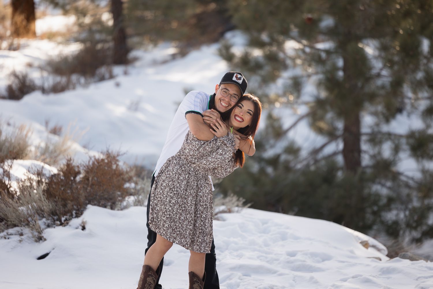 Two people embrace in a romantic pose against a snowy winter landscape with pine trees in the background.
