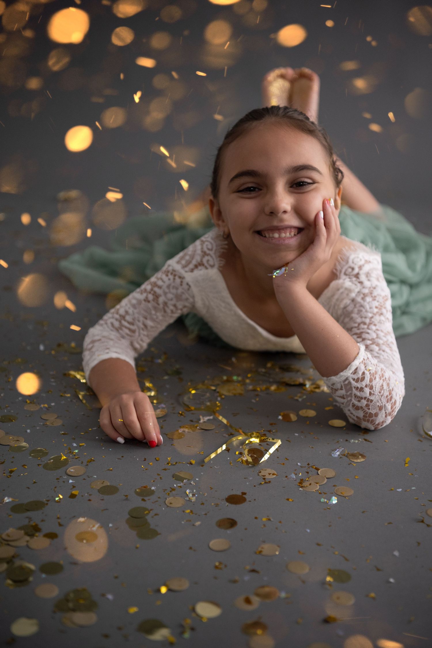 Young dancer in white lace leotard lies on floor surrounded by sparkling gold confetti and bokeh lights.