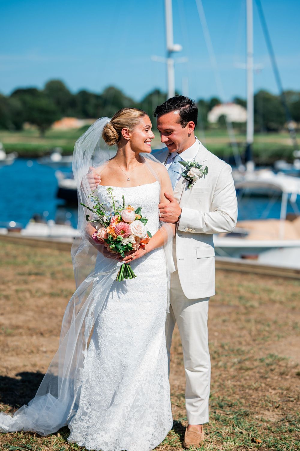 Newlywed couple embraces near a marina on their wedding day with boats and blue water in the background.
