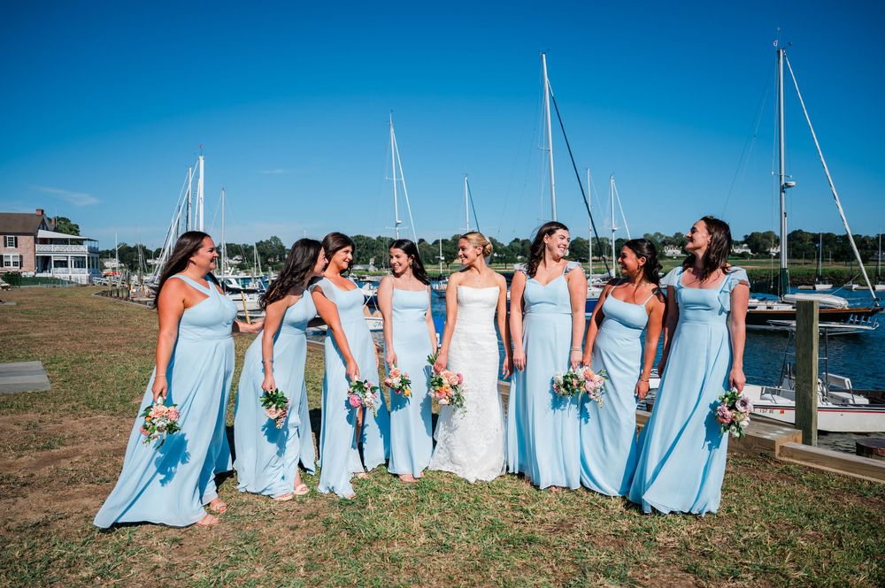 Bridesmaids in light blue dresses sit together by a marina with boats in the background on a sunny day.