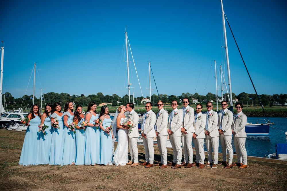 Wedding party in light blue and white attire stands lined up along a scenic waterfront with boats and blue sky.