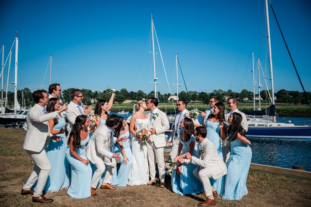 Wedding party in light blue and white attire celebrates on a dock with boats and water in the background.