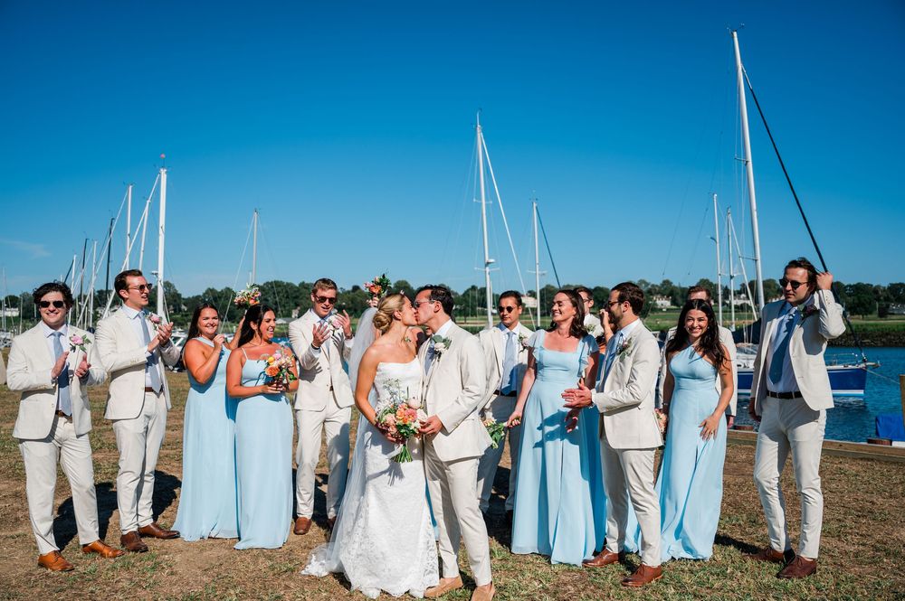 Wedding party in light blue and white attire poses along scenic waterfront marina on sunny summer day.