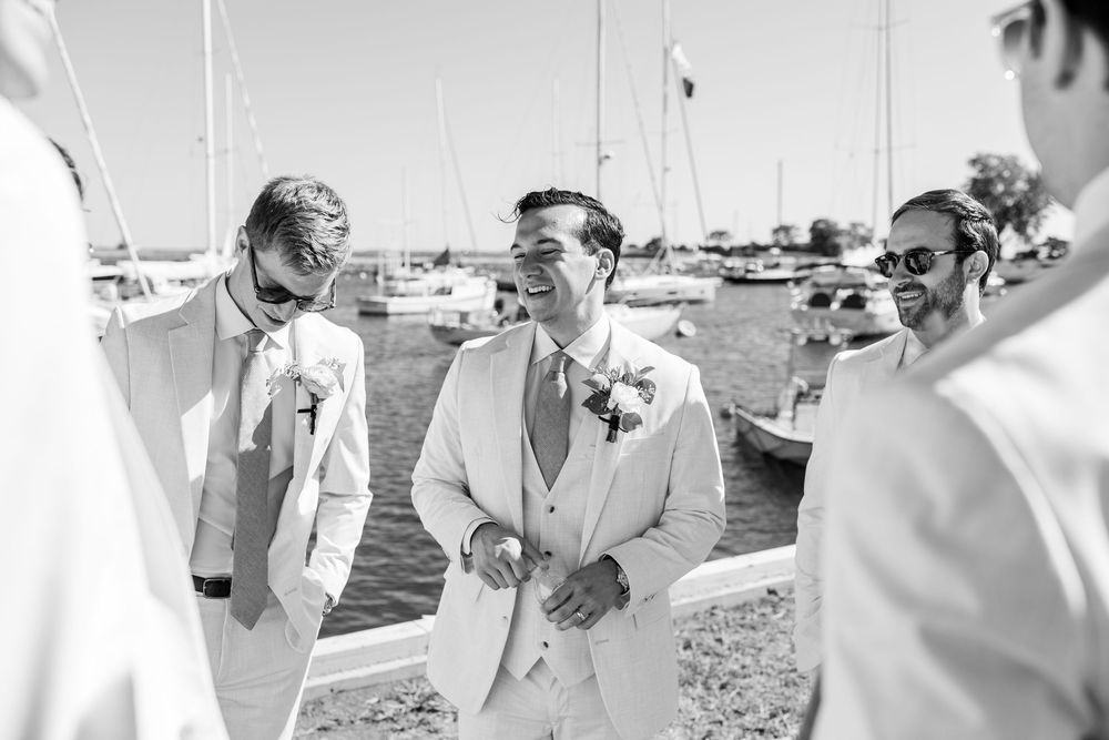 A group of people in light colored suits share a candid moment while standing near a marina with boats in the background.