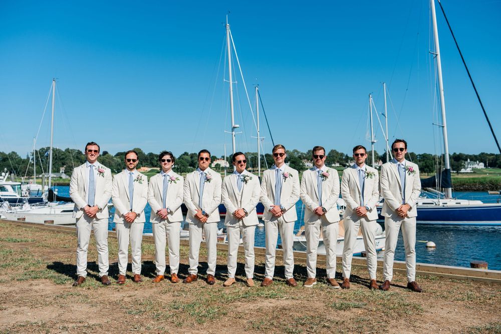 Wedding party in white attire poses in a line against a scenic marina backdrop with sailboats and blue sky.