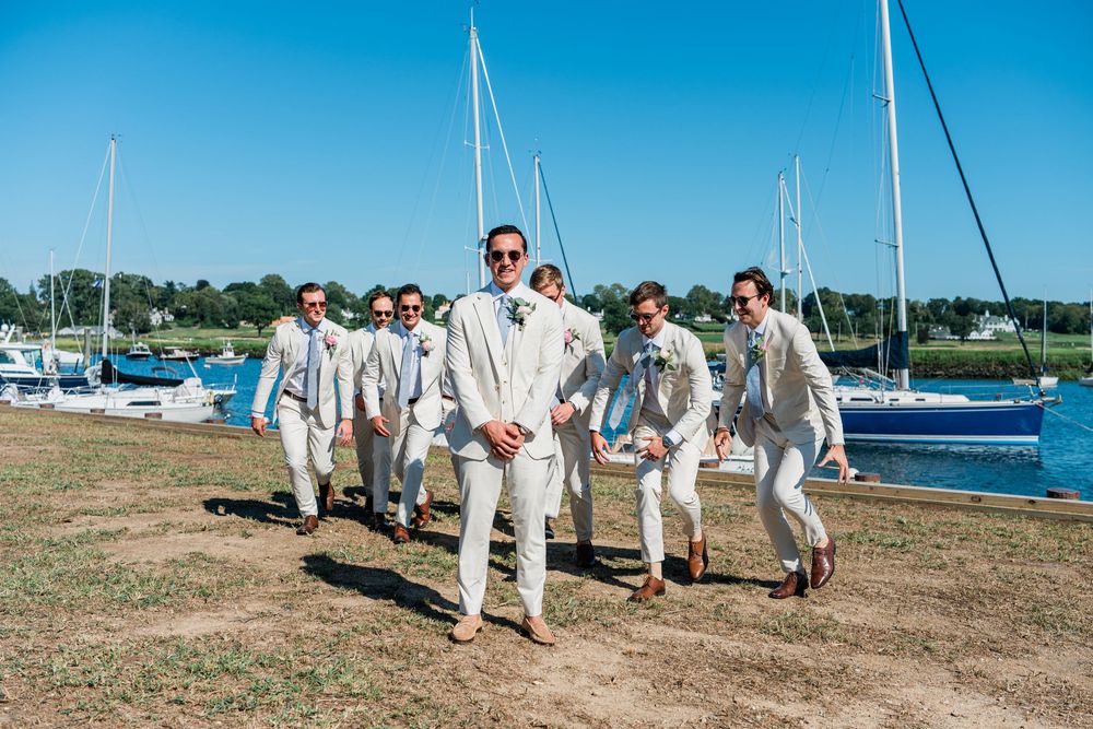 Wedding group stands on grassy shoreline with sailboats and blue water in background.