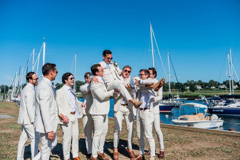 Wedding party in white outfits stands together on a grassy area near a marina with boats in the background.