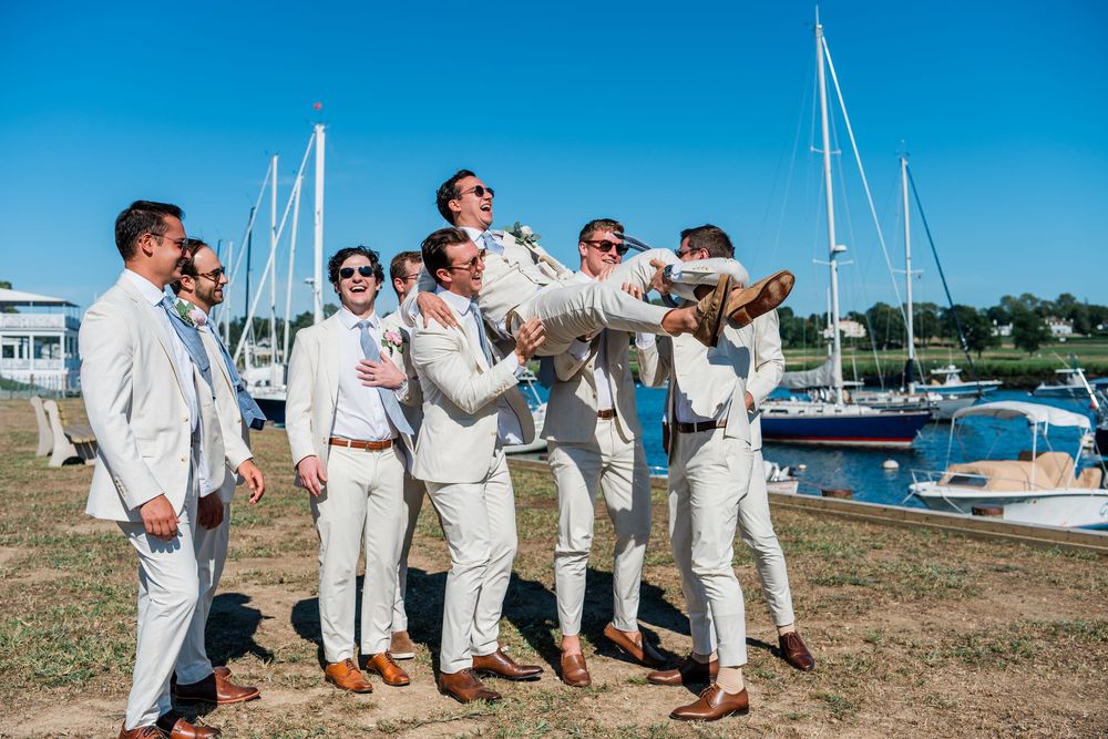 Wedding party in white suits poses playfully near a marina with sailboats and blue sky in the background.