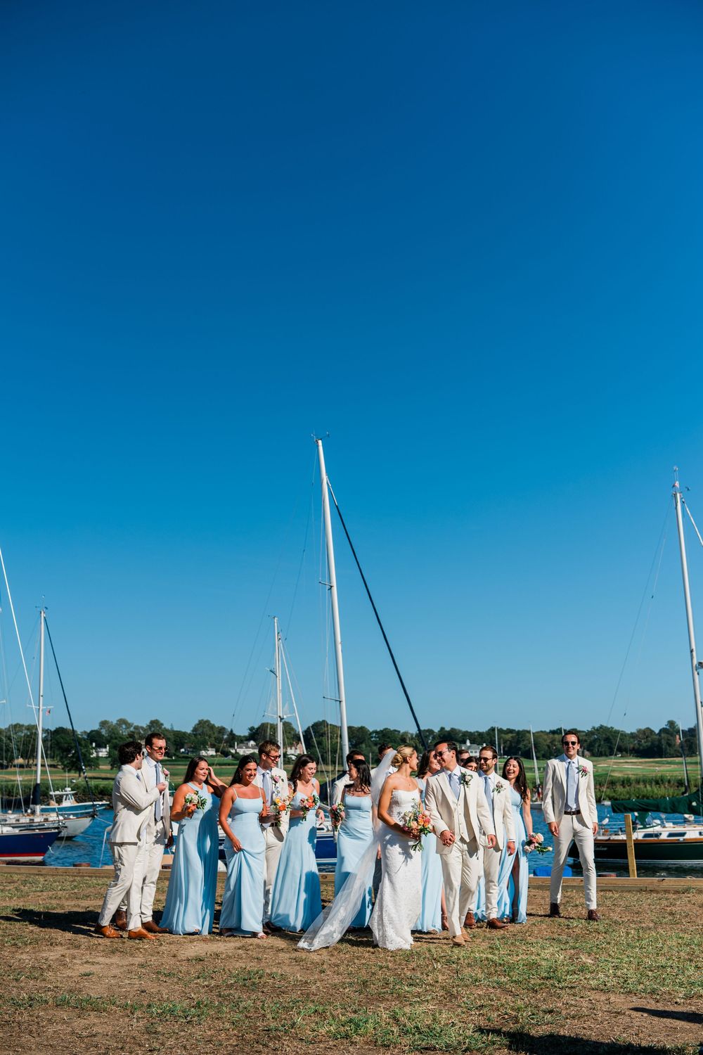 Wedding party in white and light blue dresses stands along a waterfront marina with sailboats in the background.