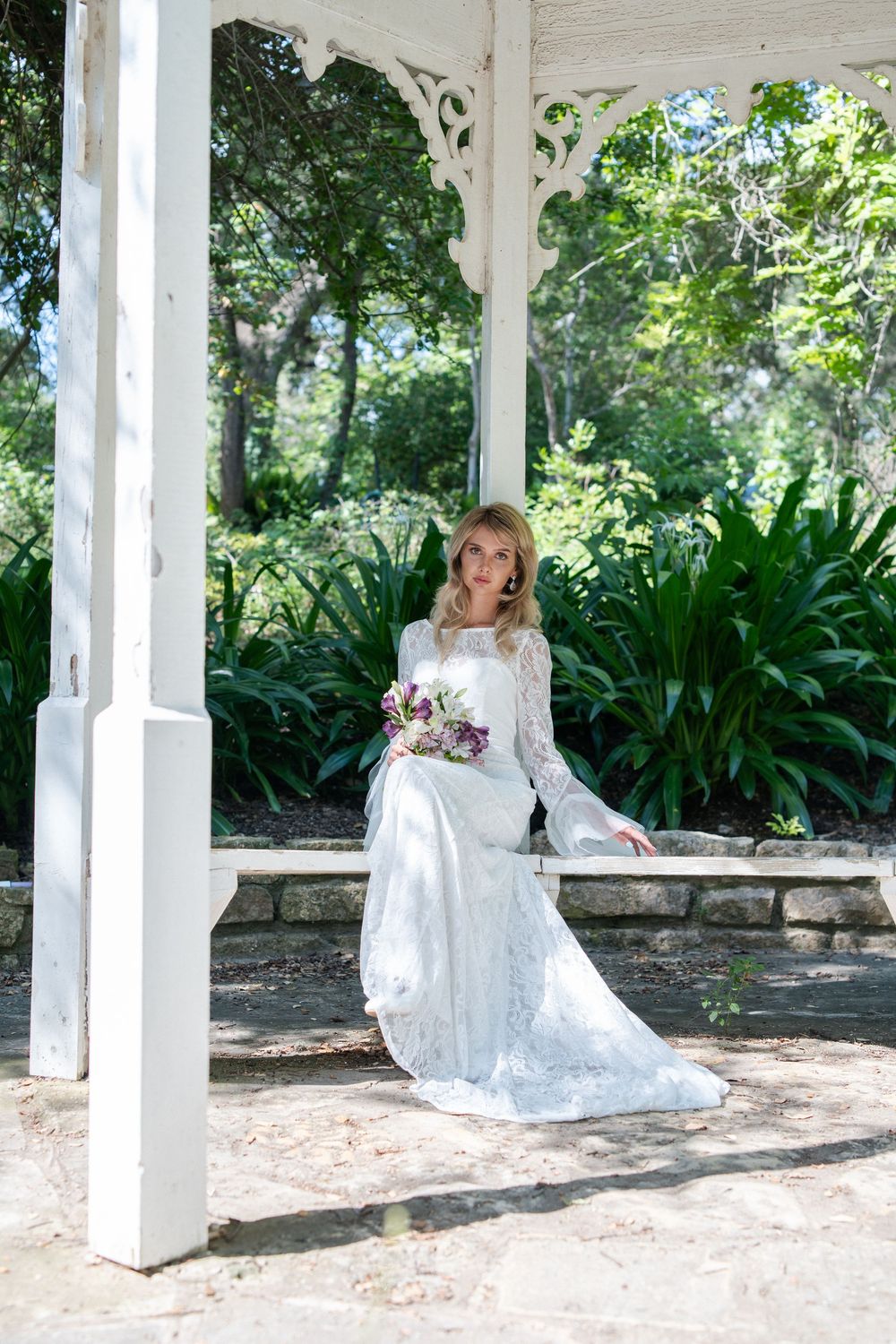 A bride in a white lace dress poses in a garden gazebo with ornate white pillars.