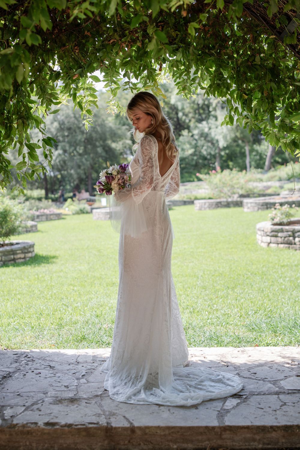 A bride in a flowing white dress poses under trees in a garden setting.
