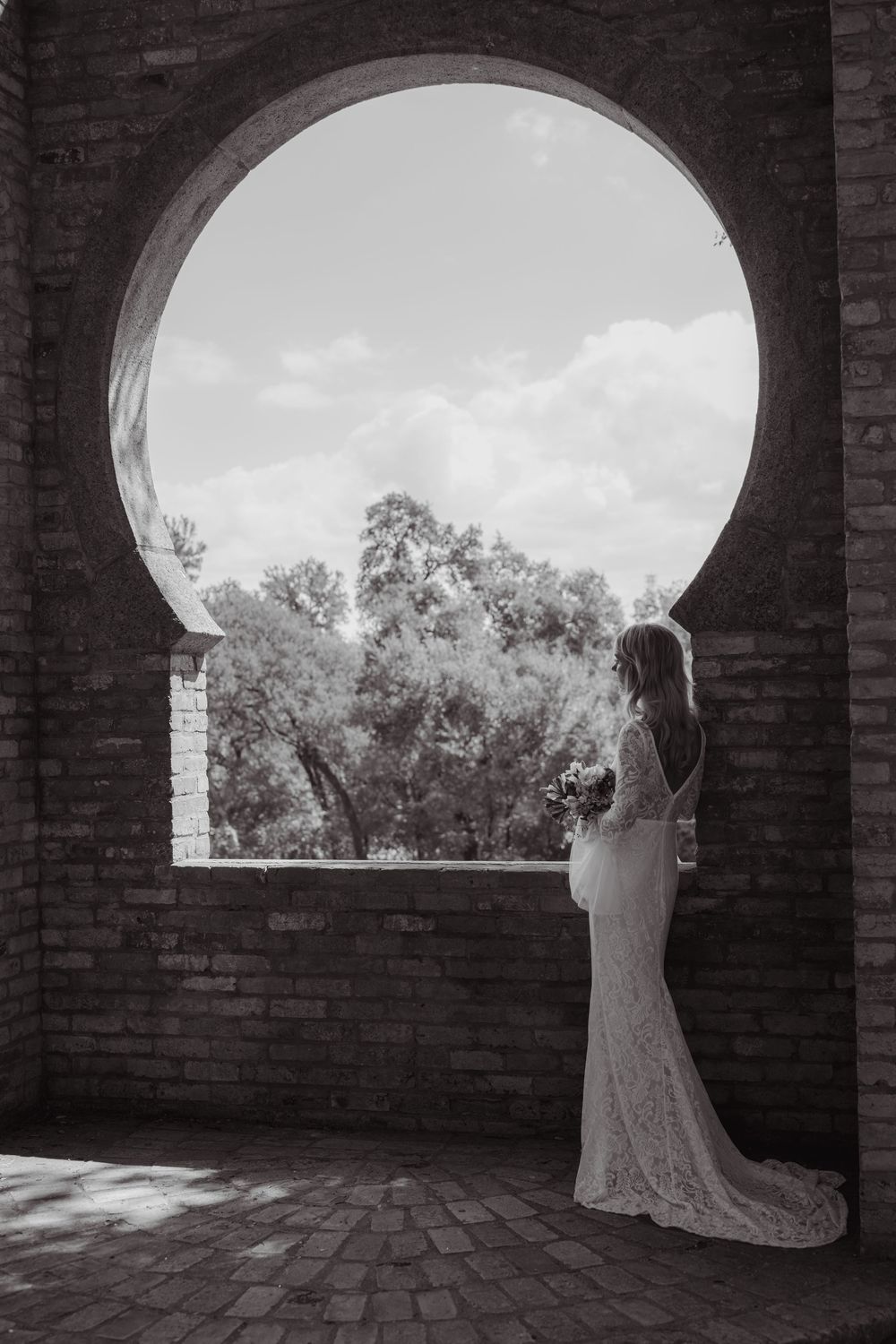 A silhouette in a wedding dress stands in an arched brick doorway overlooking a garden landscape.