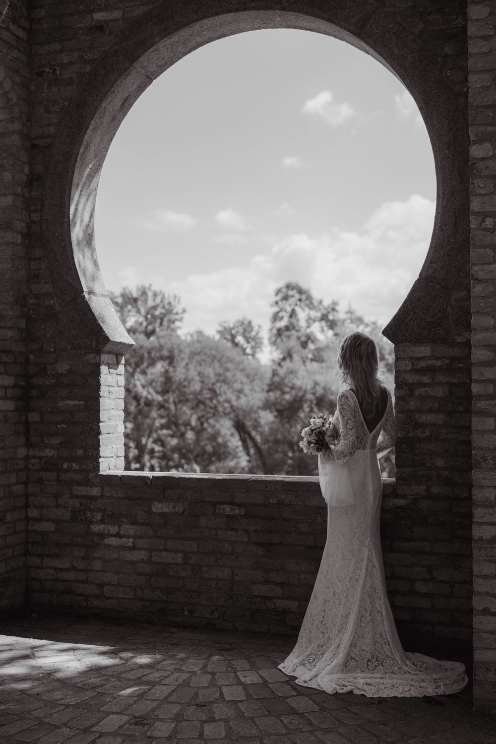 A bride poses in an arched brick window frame with a scenic garden backdrop.