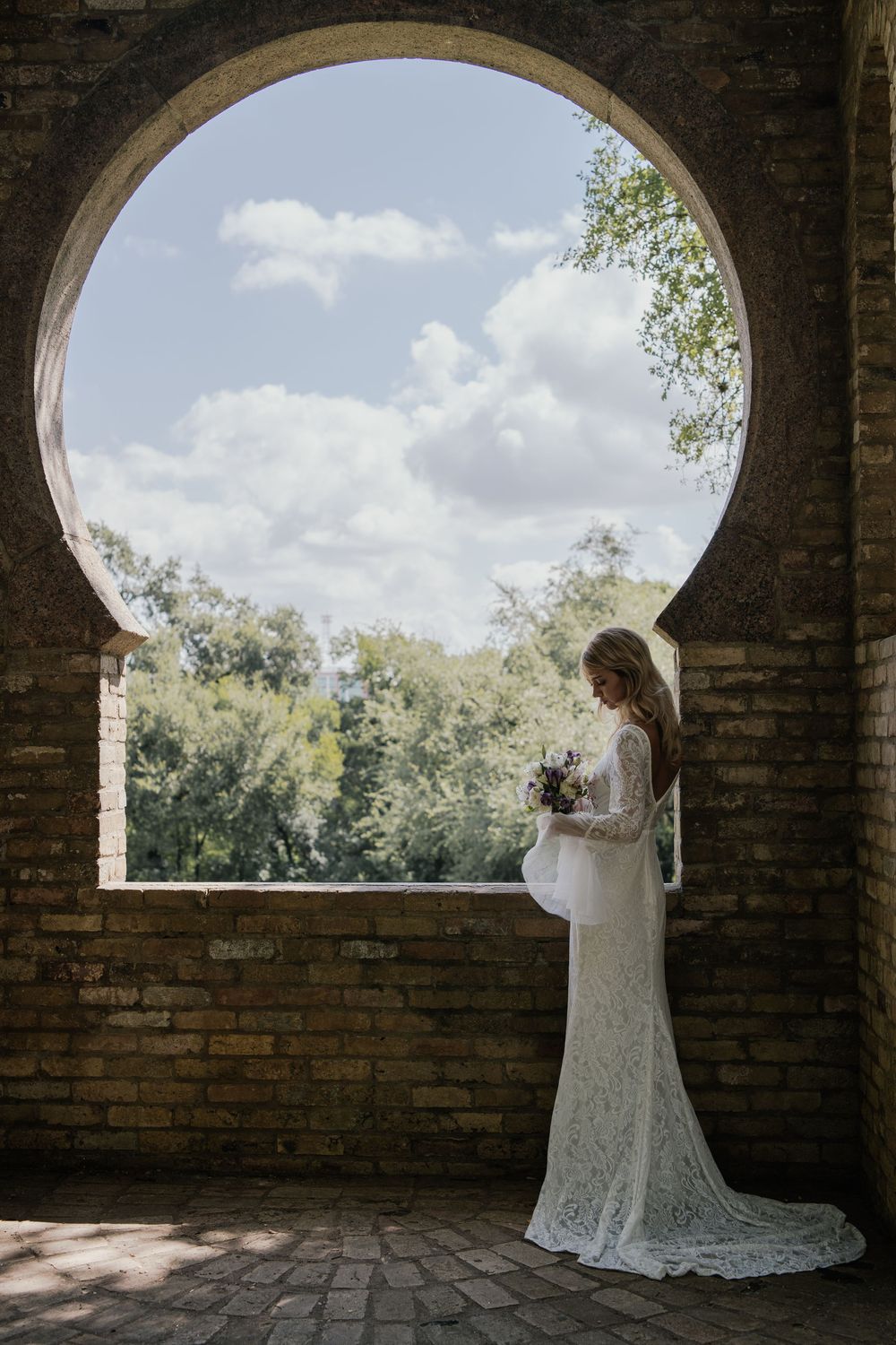 Elegant bride poses in a lace gown under arched brick windows with natural sunlight streaming through.