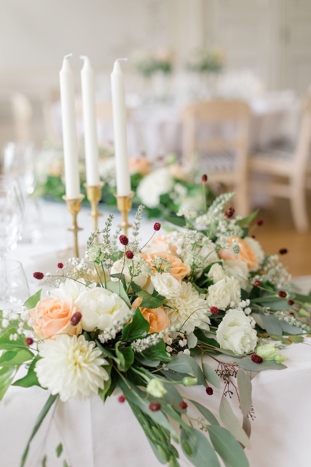 Elegant wedding centerpiece with white dahlias, peach roses, and greenery arranged with tall white taper candles.