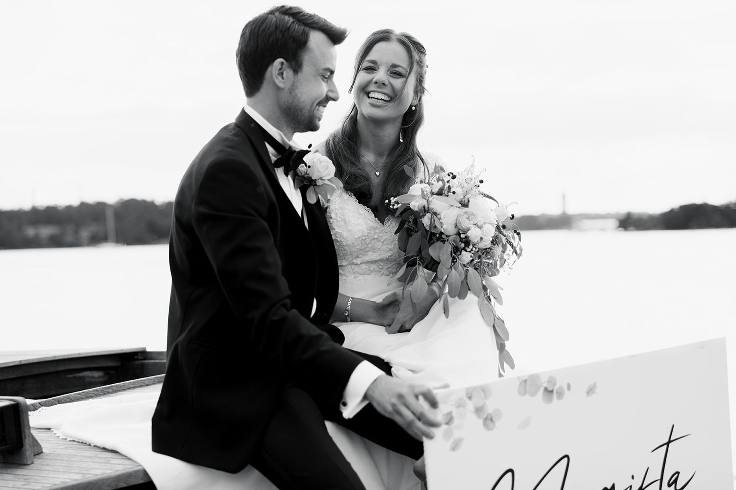 A wedding couple stands at the bow of a vintage wooden boat on a serene lake during sunset.