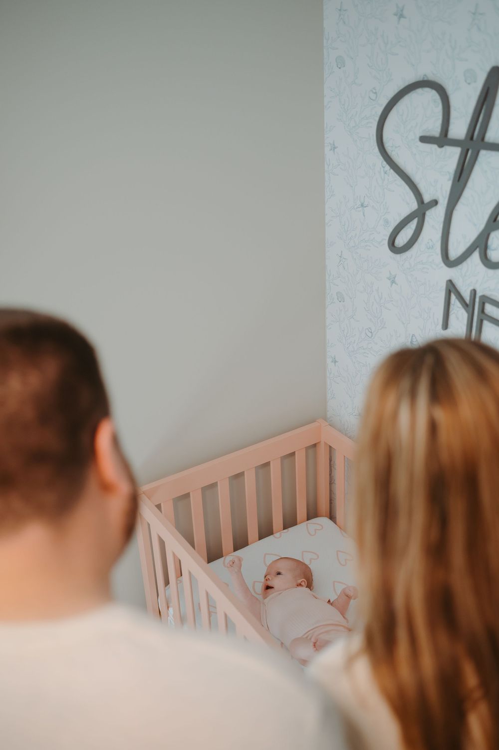 Parents lovingly watch over their sleeping baby in a wooden crib under soft nursery lighting.