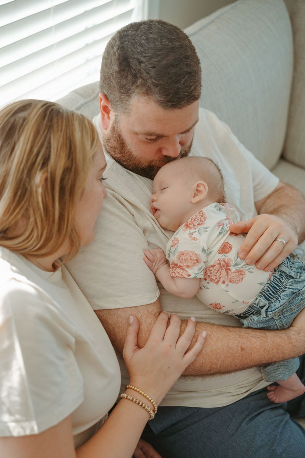 Tender family moment showing parents cuddling with small child on white couch in soft natural light.