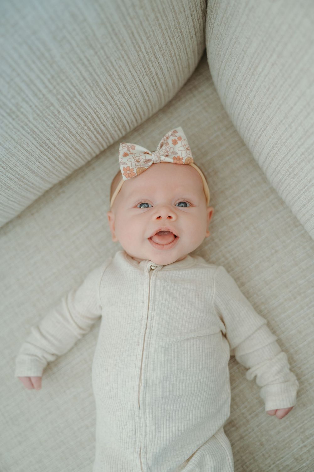 Baby wearing white ribbed onesie and peach floral bow headband smiles while lying on light colored couch.