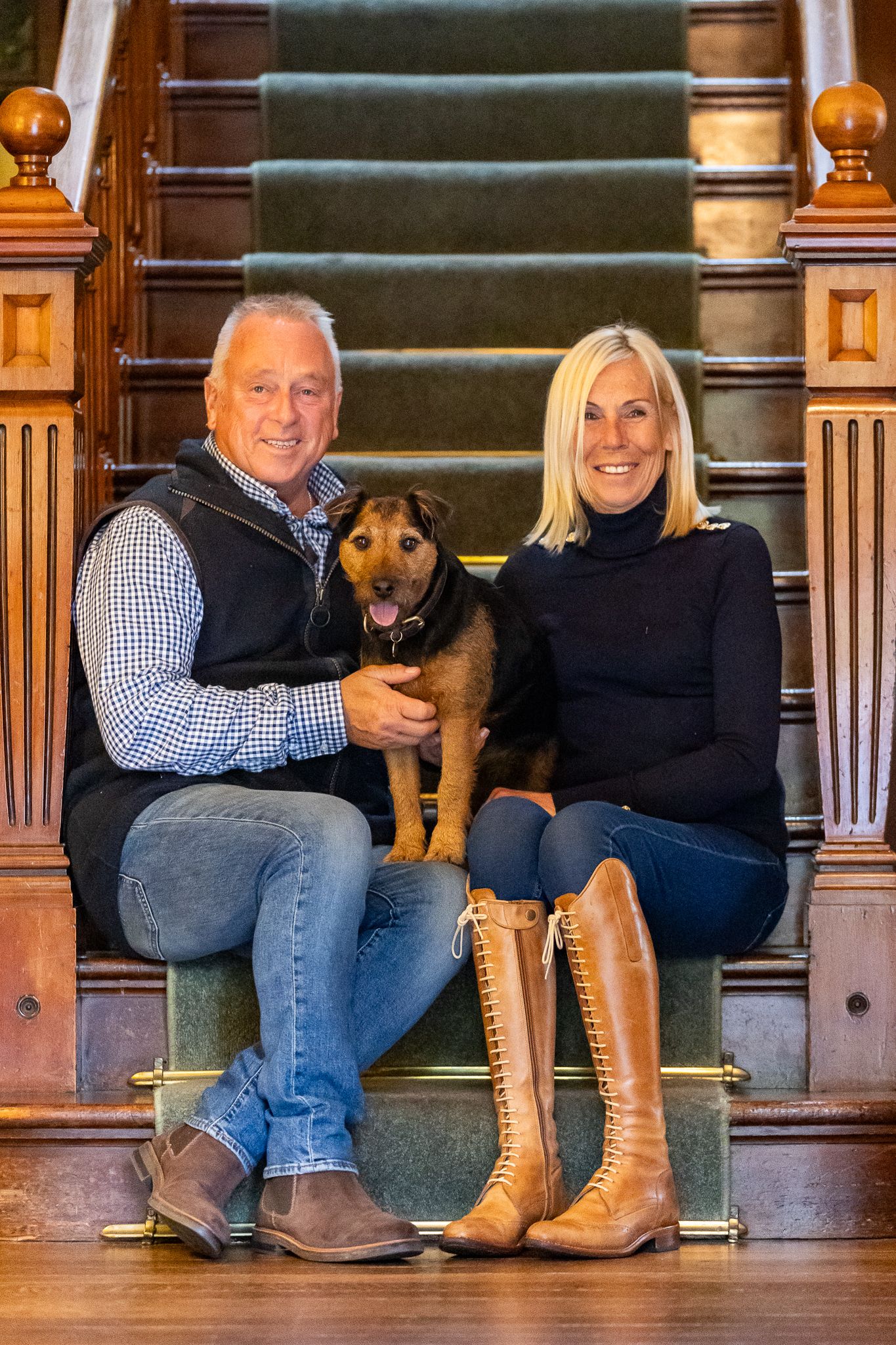 Couple sitting on elegant wooden staircase with green carpet with their brown rescue dog in cozy home setting.