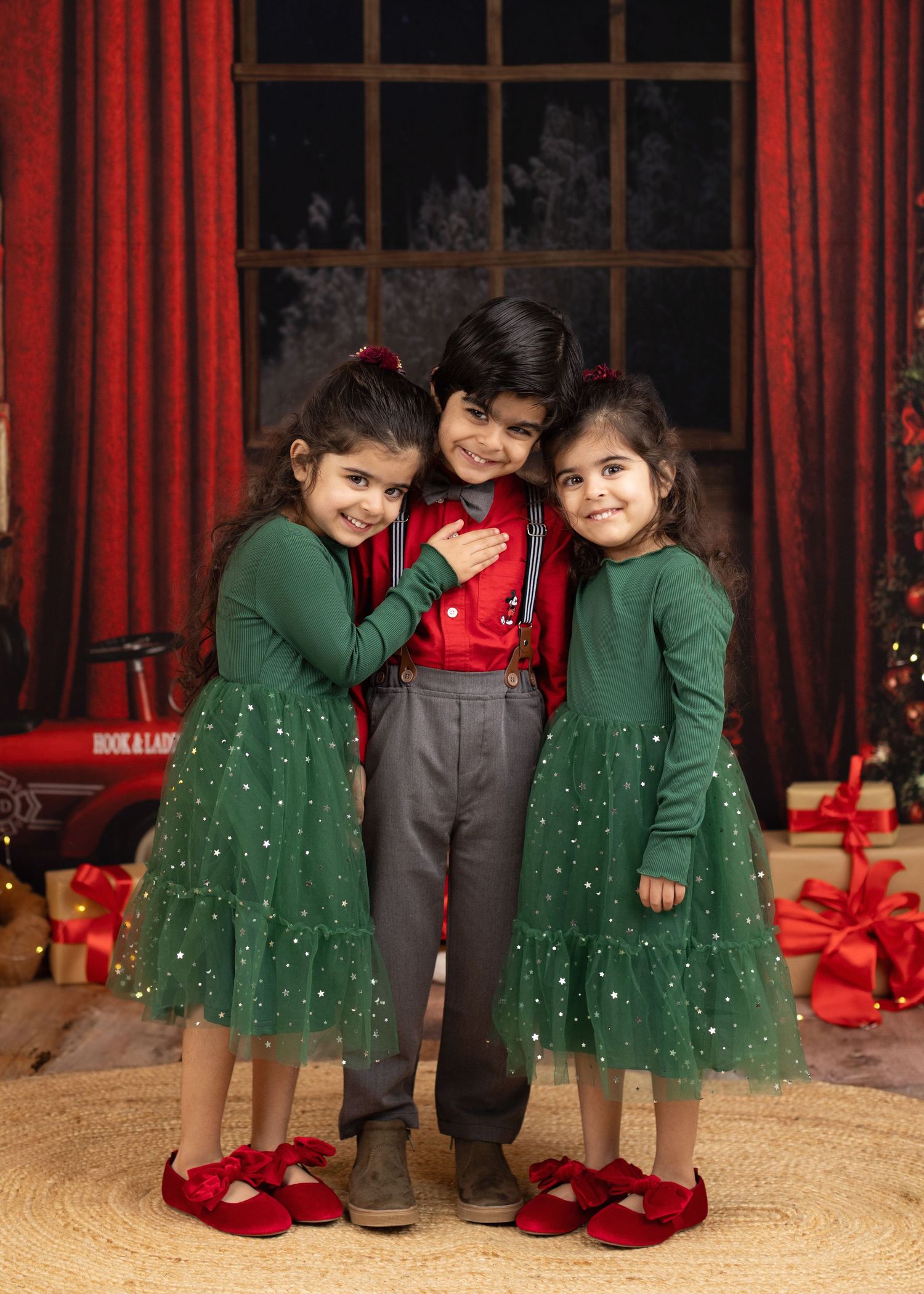 Three children in festive holiday attire pose in front of red curtains with Christmas decorations and wrapped gifts.