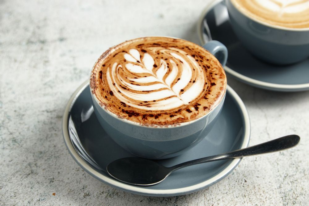 A steaming hot cappuccino with intricate latte art design served in a dark gray ceramic cup and saucer on concrete surface.