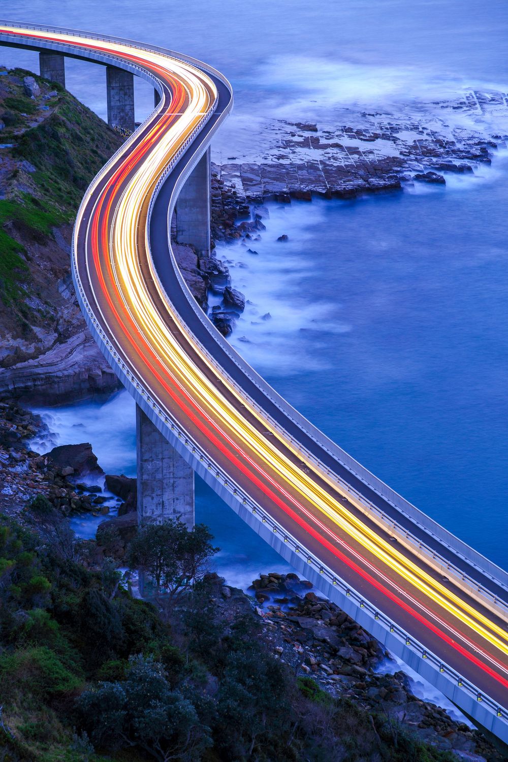 Long exposure photo of car light trails along a coastal highway curving around the ocean at dusk with blue water below.
