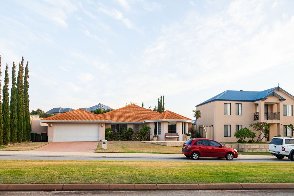 Row of residential houses with terra cotta roofs and cypress trees line a suburban street against blue sky.