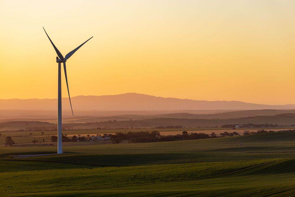 Wind turbines stand tall against a golden sunset sky on rolling green hills with mountains in the distance.