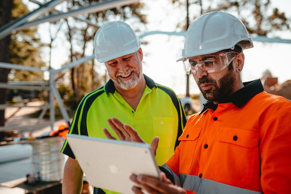 Construction workers in hard hats and safety gear review building plans together at a worksite during sunset.