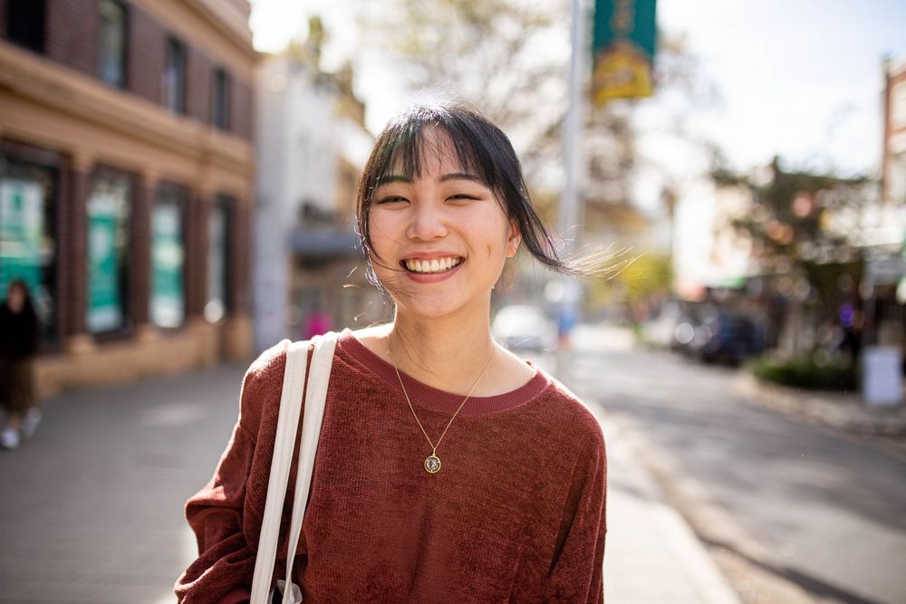 A person in a burgundy sweater smiles warmly on a sunny street with storefronts in the background.