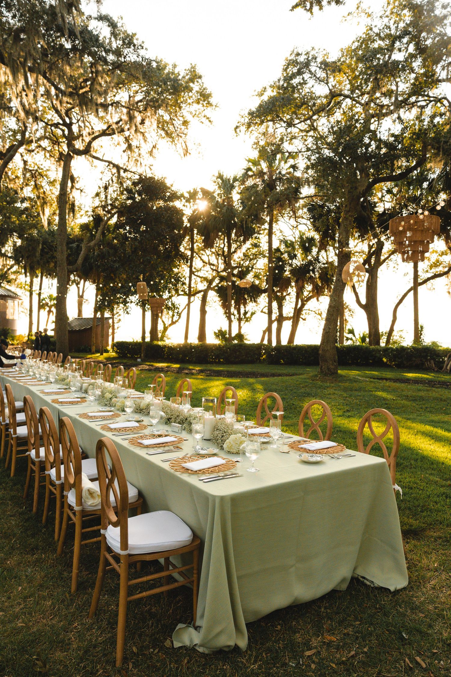 Elegant outdoor dining table setup with white linens and wooden chairs under oak trees at sunset.